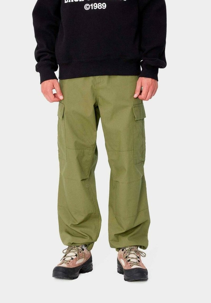 Pantalones cargo verde oliva con múltiples bolsillos laterales, diseño de pierna recta y rodillas reforzadas. Llevado con zapatos de senderismo color crema y rosa.