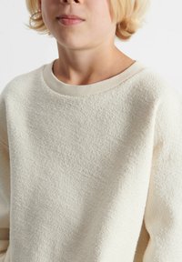 Krämsfärgad sweatshirt med en strukturerad, mjuk yta. Har rund halsringning och ribbat avslut. Utomhusbelysning, minimala synliga detaljer.