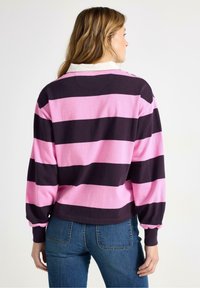 Kvinde med langt lyst brunt hår iført en pink og sort bredstribet langærmet skjorte og blå jeans, set bagfra.