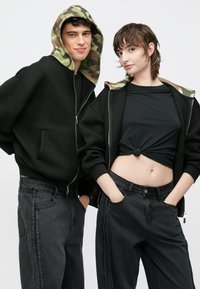 Fekete cipzáros kapucnis pulóverek terepmintás béléssel, fekete magasderekú farmerekkel kombinálva. A női modell fekete, megkötött felsőt visel, amely kibontja a hasát.