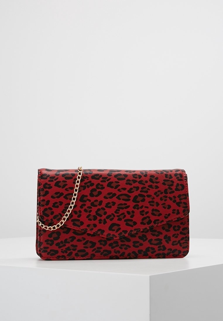 Pochette rouge avec imprimé léopard noir et bandoulière chaîne dorée sur une surface blanche sur fond gris clair uni.