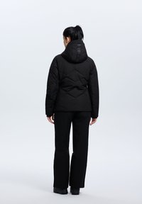 Svart pufferjacka med huva, med en quiltad design. Kombinerad med svarta byxor och stiliga skor. Minimalistisk, funktionell stil.