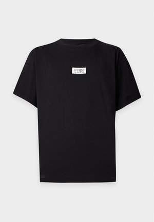 Ung mand iført en sort t-shirt, sort plisseret nederdel med bælte, brede sorte bukser og sorte sko, står mod en ensfarvet baggrund.