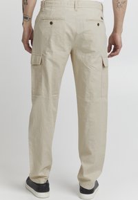 Pantalons cargo beiges avec poches latérales, poches arrière à bouton, tissu léger et coupe décontractée, associés à des baskets noires.