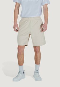 Pantaloni atletici beige con cintura elastica, dotati di un dettaglio del logo sulla gamba. Abbinati a una maglietta bianca e sneaker bianche.