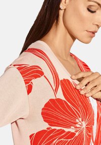 Blumen-Cardigan in zartem Rosa mit großen roten Blumenmustern, mit einem V-Ausschnitt und weichem Griff. Detaillierte Steppnähte entlang des Ausschnitts.