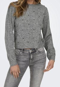 Pull gris tricoté avec un motif chevron, orné de petites perles noires. Comprend des manches longues et un ourlet court. Associé à un jean gris.