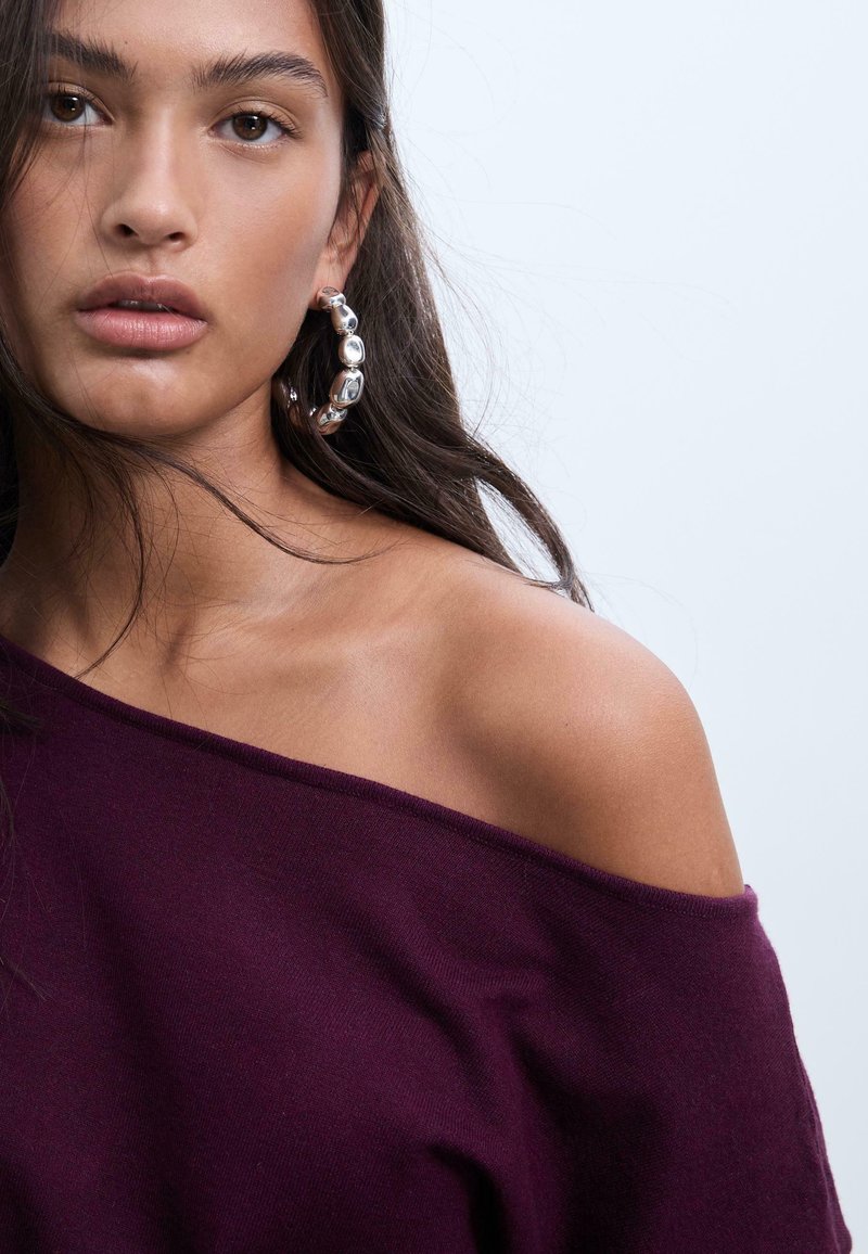 Pull moelleux bordeaux à épaules dénudées. Il présente un col lisse et une boucle d'oreille en argent avec un design arrondi.