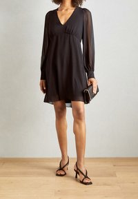Schwarzes knielanges Kleid mit V-Ausschnitt, langen transparenten Ärmeln und geraffter Taille, kombiniert mit schwarzen Riemchenabsätzen und einer kleinen Clutch.