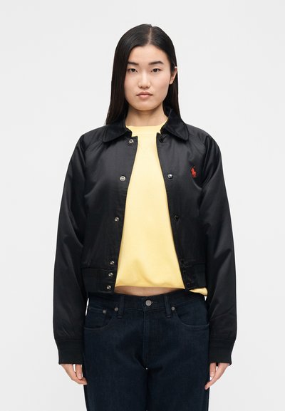 Čierna krátka bunda z hladkej látky so zapínaním na patentky, špicatým golierom a červeným logom. Nosí sa cez žltú mikinu.