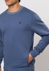 Modra dukserica iz gladkega blaga, z okroglim izrezom s turkiznim robom, stranskimi žepi in vezenim logotipom na prsih.