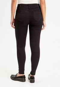 Sorte slim-fit leggings med en glat tekstur, der har dekorative knapper ved taljen og to baglommer, parret med glinsende sorte sko.