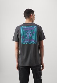 Camiseta de algodón gris con un diseño gráfico colorido en la parte posterior, que incluye rayas diagonales y una ilustración de una cara geométrica.
