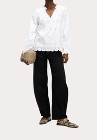 Blouse blanche en dentelle avec un col et des poignets à volants, assortie à un pantalon noir à jambes larges et un sac à main beige ; elle est complétée par des ballerines à motifs.