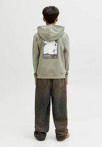 Osoba v svetlozelenom mikinovom svete s kapucňou a čiernobielym horským grafikom na chrbte, skombinovaná s voľnými tmavými nohavicami a béžovými topánkami.