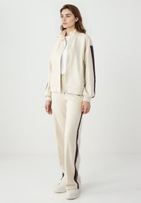 Beige Zip-Up-Jacke mit hohem Kragen, weichem Stoff und seitlichen Streifen. Passende beige Hose mit seitlichen Streifen und weißen Sneakers.