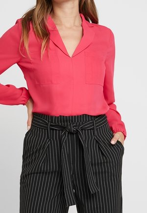 Mujer que lleva una blusa de manga larga de color rosa brillante metida en pantalones de rayas negros de talle alto con un cinturón de lazo a juego.
