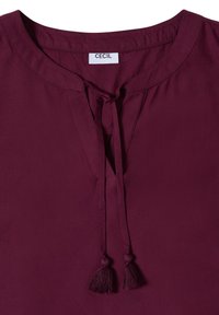 Blouse bordeaux à encolure fendue, cordons terminés par des pompons et une étiquette "Cecil" au col.