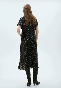 Schwarze Bluse mit Polka-Dots und lockerem Schnitt, kombiniert mit einem langen schwarzen Rock. Weicher Stoff, knielanges Design, getragen mit knöchelhohen braunen Stiefeln.