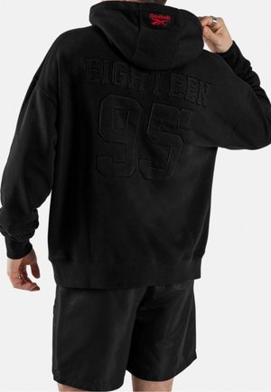 Persona che indossa una felpa nera con cappuccio con "EIGHTEEN 95" impresso sulla schiena e logo rosso Reebok sul cappuccio, abbinata a pantaloncini neri.