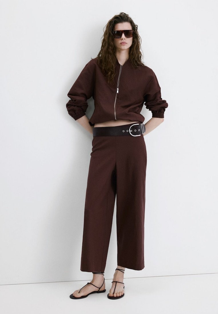 Femme portant une veste courte zippée marron foncé et un pantalon à jambes larges, ceinture noire, grandes lunettes de soleil, et sandales plates noires à brides, debout contre un mur blanc.