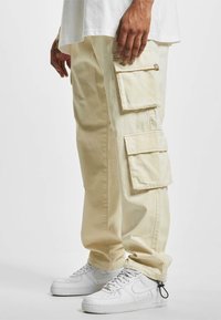 Pantalones cargo beige con múltiples bolsillos laterales, hechos de algodón, con un ajuste relajado y un dobladillo ajustado, combinados con zapatillas blancas.