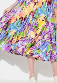 Abito floreale multicolore con balze arricciate, caratterizzato da vivaci motivi di viola, blu, giallo e verde. Bottoni visibili lungo la parte frontale.