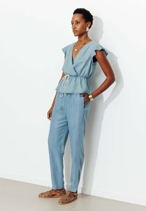 Femme appuyée contre un mur blanc, portant un haut cache-cœur sans manches bleu clair, un pantalon assorti, des bijoux en or et des sandales à imprimé léopard.