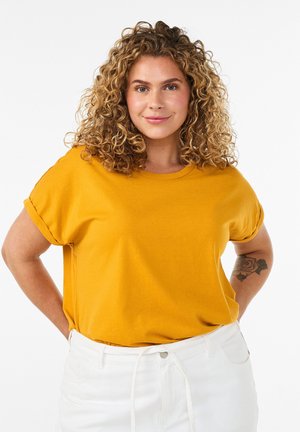 Kvinde med krøllet hår iført en sennepsgul t-shirt og hvide bukser, smilende, med en rosetatovering synlig på venstre underarm.