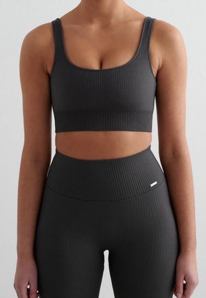 Frau trägt einen schwarzen gerippten Sport-BH und passende hoch taillierte Leggings und steht vor einem einfachen hellen Hintergrund.