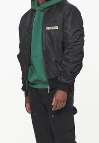Giacca bomber nera con polsini a costine, chiusura con zip e tasche laterali. Abbinata a un hoodie verde, con tasca frontale e cordino.