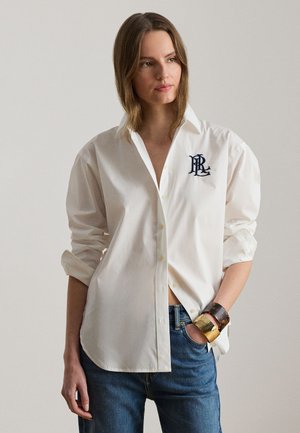 Une femme porte une chemise blanche boutonnée ornée d’un monogramme, aux manches retroussées, un jean bleu et un large bracelet manchette doré.