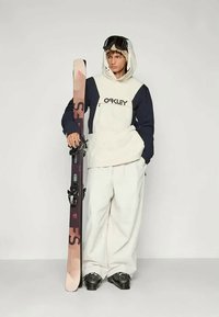 Snowboarder iført cremefarvet og marineblå hættetrøje med trykt logo, hvide bukser, sorte støvler, der holder på et farverigt ski med bindinger og skibriller.