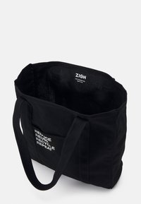 Sac fourre-tout en coton noir avec bretelles, poche avant avec le texte "RÉDUIRE RÉUTILISER RECYCLER RÉPÉTER", étiquette de marque "ZIGN" à l'intérieur.