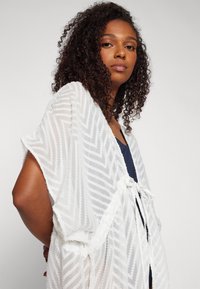 Kimono bianco aperto davanti con un motivo a zigzag testurizzato, caratterizzato da maniche corte e chiusura legata. Tessuto leggero e semitrasparente.