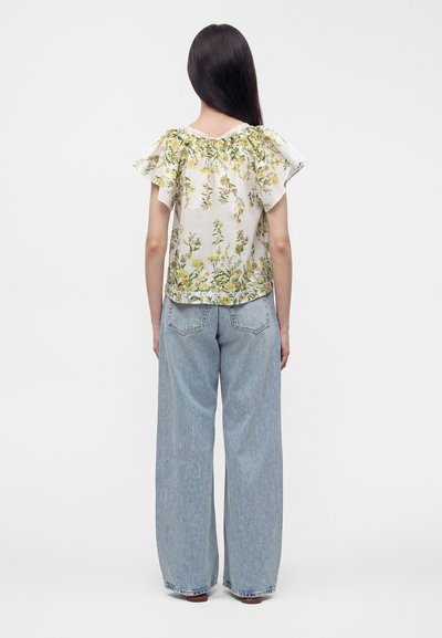 Femme aux longs cheveux foncés portant un chemisier blanc à fleurs à manches courtes avec des volants et un jean bleu clair ample, face à un fond blanc, de dos.