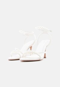 Λευκά σανδάλια με ψηλό τακούνι και λουράκια στον αστράγαλο, διακοσμημένα με μεγάλες και μικρές λευκές μαργαριταρένιες χάντρες.