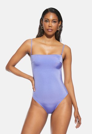 Kobieta ubrana w dopasowany, lawendowy jednoczęściowy kostium kąpielowy z cienkimi ramiączkami, pozująca z jedną ręką na biodrze na białym tle.