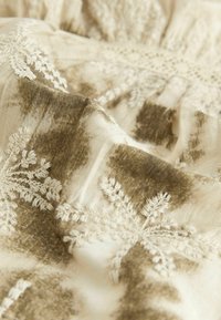 Beige stof med indviklet brunt blomsterbroderi og subtile teksturforskelle, der har delikat syning og et blødt, flydende design.