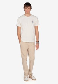 Camiseta de algodón color beige claro con un pequeño diseño gráfico colorido en el lado izquierdo del pecho, combinada con pantalones beige claros y zapatos casuales.