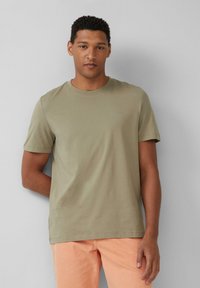 Oliwkowa bawełniana koszulka z krótkim rękawem, okrągłym dekoltem i subtelnym logo na piersi. W zestawieniu z jasnopomarańczowymi spodniami.