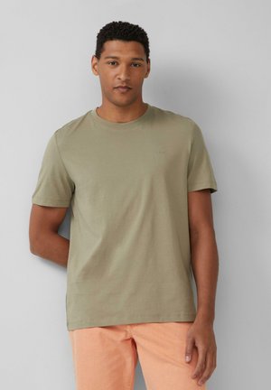 Olijfgroene katoenen T-shirt met korte mouwen, ronde hals en een subtiele logo op de borst. Gecombineerd met licht perzikkleurige broeken.