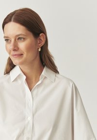 Camicia bianca con colletto, dal taglio rilassato e in morbido cotone. Il modello indossa orecchini a cerchio dorati e volge leggermente a sinistra.