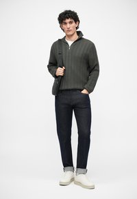 Σκούρο πράσινο καπιτονέ ζακέτα με φερμουάρ, σκούρες τζιν παντελόνες, λευκά αθλητικά παπούτσια και μια μαύρη τσάντα ώμου. Καθημερινό ντύσιμο με δομημένο σχεδιασμό.