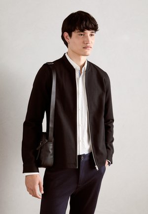 Veste noire zippée avec col montant, portée sur une chemise blanche à boutons. Présente une texture lisse et un sac noir porté en bandoulière.