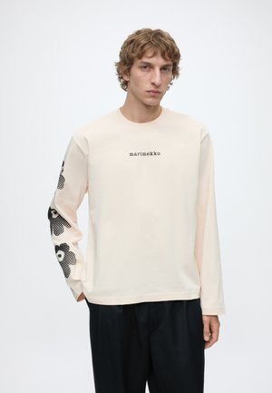 Jonge man draagt een lichtbeige shirt met lange mouwen met de tekst "marimekko" op de borst en zwarte bloemmotieven op de linkermouw.