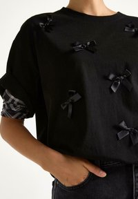 Person iført en sort skjorte med seks små sorte satinbuer, oprevne ærmer der afslører et sort-hvidt mønstret lag nedenunder.