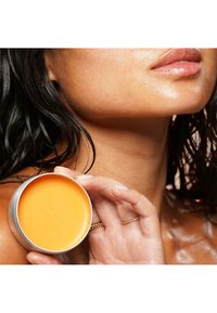 Rundes silbernes Döschen mit einem leuchtend orangefarbenen Produkt, das in einer Hand gehalten wird, daneben ein Nahaufnahme der Haut mit glatter Textur und feuchtem Aussehen.