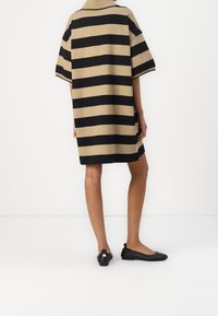 Randig, oversized klänning i beige och svart, med hög halsringning och korta ärmar. Bärs med svarta ballerina-skor, som visar på en slät textur.