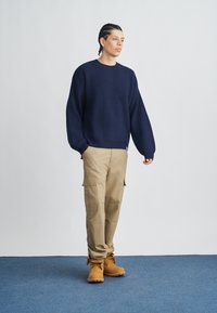 Maglione lavorato a maglia blu navy con texture a coste, pantaloni cargo beige e stivali color miele. Mani in tasca, in piedi su un tappeto blu contro uno sfondo chiaro.